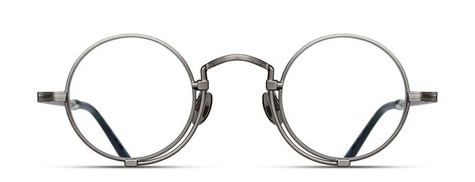 Optical glasses frames are pictured, displaying a frontal view. The glasses feature a round lens shape outlined in antique silver and a M + N engraving along the rim and temple. 