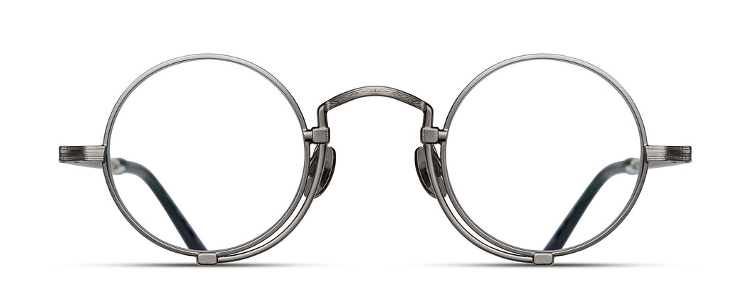 Optical glasses frames are pictured, displaying a frontal view. The glasses feature a round lens shape outlined in antique silver and a M + N engraving along the rim and temple. 